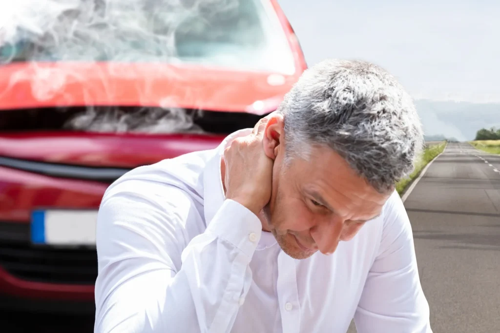 A man holding his neck with his car crashed in the background.
