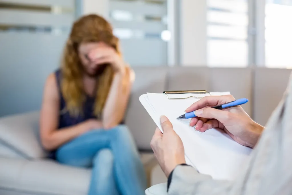 An upset woman sitting on the couch talking to her doctor who is taking notes.