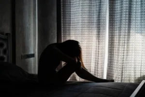 A woman sits on a bed with her head down and arms wrapped around her knees, silhouetted against a curtained window, conveying sadness or emotional distress.