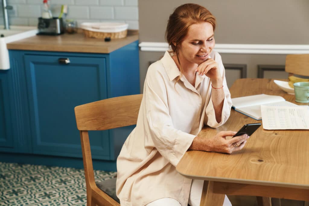 A woman at her kitchen table looking at her phone beside paperwork and a notebook