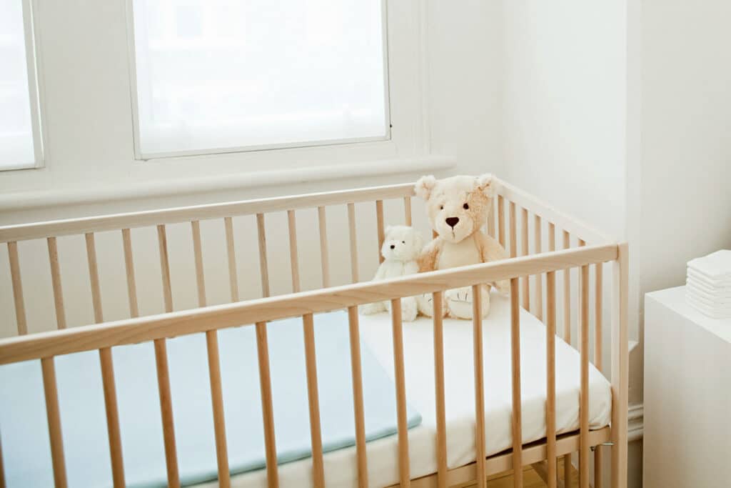 An empty wooden crib beside a window in a quiet nursery, lit by soft daylight