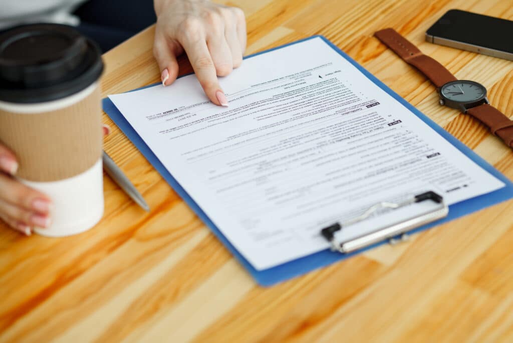 A hand pointing at a printed form on a clipboard at a kitchen table, coffee nearby