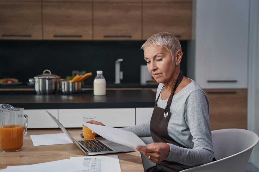A woman reviewing a printed document beside an open laptop in her kitchen