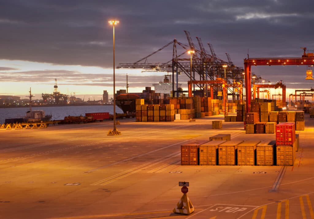 Stacked shipping containers in a freight terminal yard at dusk, with port cranes and a ship in the background