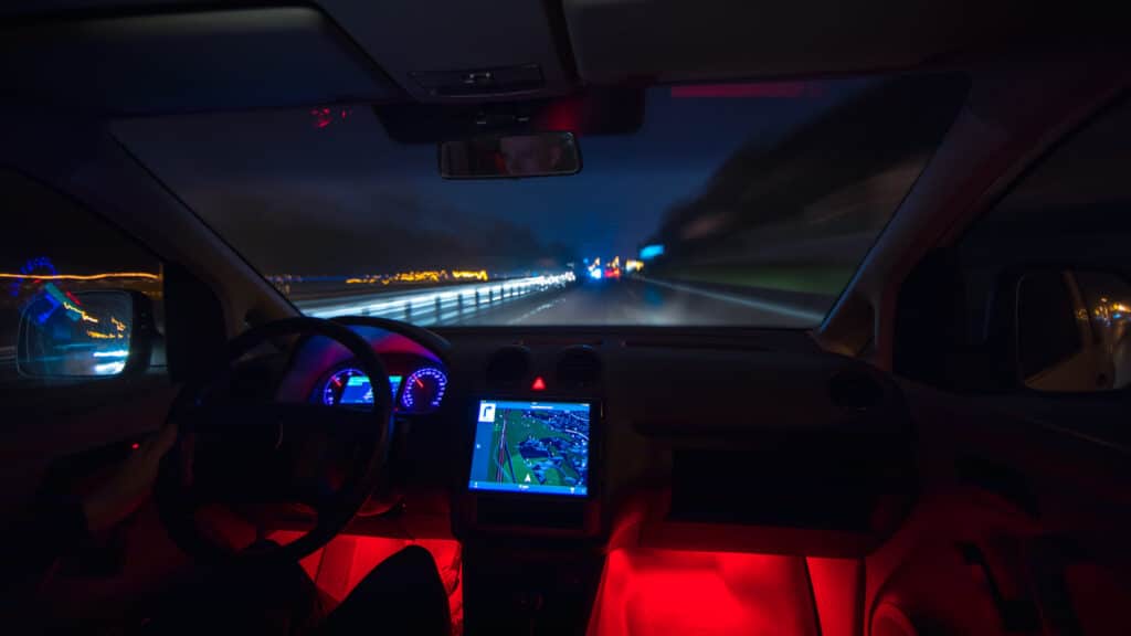 A car interior at night, dashboard lit red, navigation screen glowing, seen through the windshield on a highway