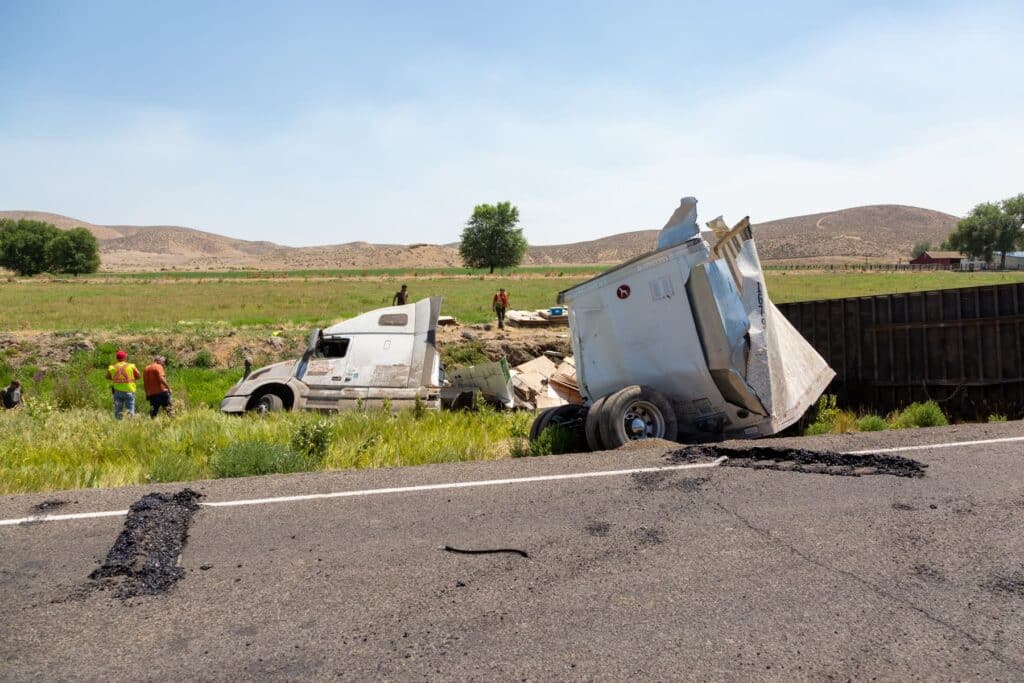 An overturned tractor-trailer resting in a grassy field beside a highway, seen from a distance