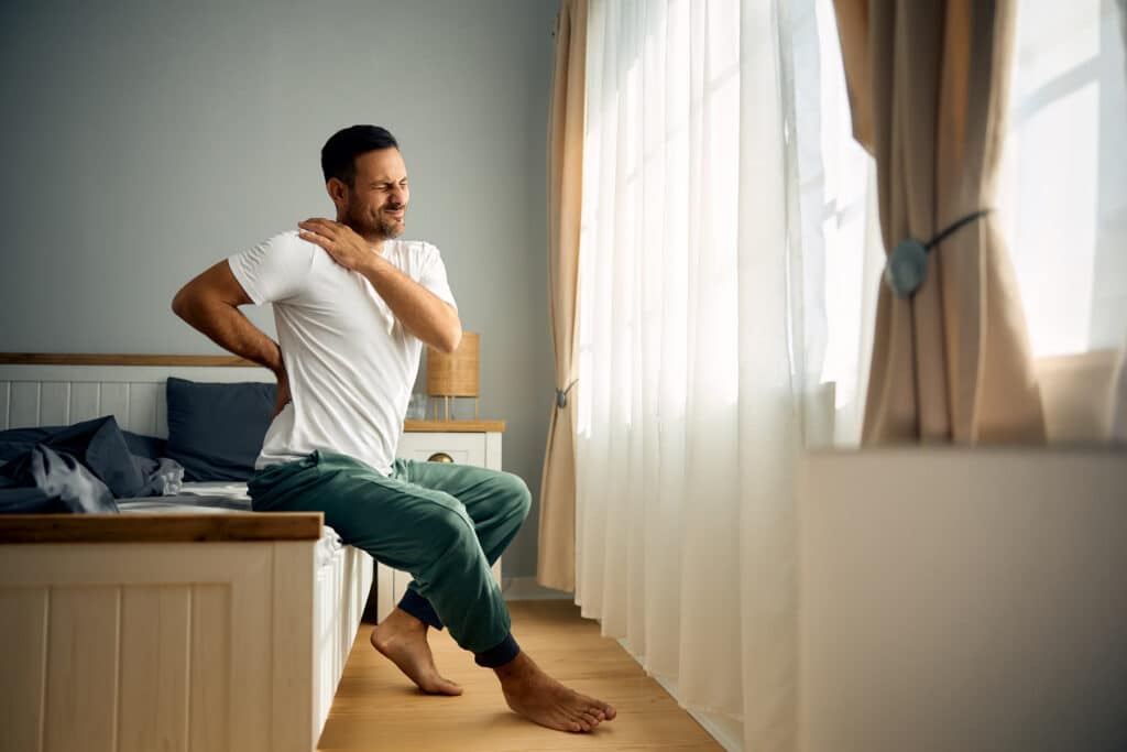 A man sitting on the edge of a bed in a bright bedroom, holding his neck and shoulder with a pained expression